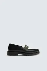 Mocasín de cuero con diseño de antifaz bicolor, combinando negro y blanco, con un detalle de tira verde oliva texturizada. Posee horma redonda y suela track gruesa de goma.