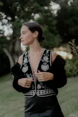 Chaleco negro con escote en V profundo, confeccionado en tela con bordados florales en hilo plateado. Presenta un diseño estructurado con ribetes plateados en los bordes y cierre frontal.