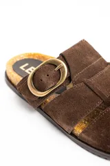 Sandalias tipo zueco de cuero gamuzado color marrón, con diseño de tiras cruzadas, puntera cerrada y una gran hebilla metálica dorada en el lateral. Cuenta con base de corcho y suela plana.