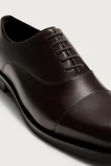 Zapato de vestir tipo oxford con puntera reforzada, confeccionado en piel de color marrón oscuro. Presenta cierre mediante cordones al tono, forro interior y plantilla de piel, y suela a contraste con tacón bajo.