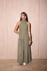 Vestido largo holgado color verde oliva, con escote halter y cuello alto con volado. Presenta un corte con fruncido a la altura de la cadera que genera un maxi volado en la falda.
