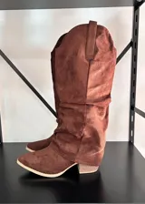 Botas texanas de caña alta en material símil gamuza color marrón, con diseño arrugado, punta fina y taco cuadrado de madera.