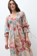 Vestido largo de manga larga, con escote en V y fruncido bajo el busto. Presenta un estampado tipo paisley en tonos rojo, azul claro y beige. El ruedo termina con un volante fruncido.