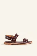 Sandalias planas de cuero charol color bordó, con tiras decoradas con tachas metálicas plateadas y suela baja.