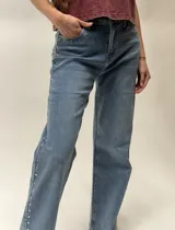 Jean de denim azul de corte recto, con detalles de tachas metálicas plateadas en los bolsillos delanteros y a lo largo de la costura exterior de la pierna derecha.