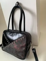 Cartera rígida negra con detalles en rojo y tachas plateadas. Tiene herrajes y cierre metálico plateado marca YKK, asa fina para llevar al hombro o en la mano y correa larga extra regulable. El interior es forrado, tiene un bolsillo con cierre enmarcado en cuero, y un segundo bolsillo plaqué.