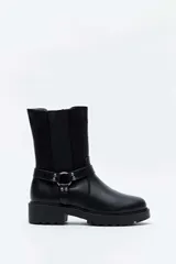 Bota alta color negro, con suela track y taco cuadrado. Presenta una tira con tachas y una argolla metálica decorativa alrededor del tobillo, además de inserciones elásticas laterales.