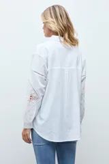Camisa blanca de corte recto con cuello clásico y cierre frontal de botones. Presenta mangas largas con detalles de encaje calado en los antebrazos y puños abotonados.