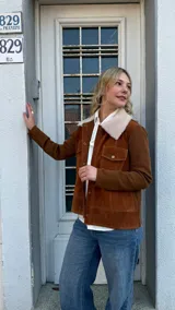 Campera de gamuza color tostado con cuello de corderito sintético en color crema. Presenta cierre frontal con botones metálicos a presión, bolsillos frontales con solapa y mangas de tejido de punto al tono.