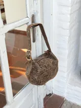 Bolso tipo hobo de cuero con acabado desgastado en tono visón. Presenta una correa ajustable con hebilla metálica y diseño de estructura redondeada.