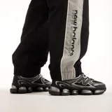 Pantalón New Balance de hombre, modelo Trackside, color negro con detalles laterales grises y logo impreso en la pierna. Cuenta con cintura elástica y diseño deportivo.