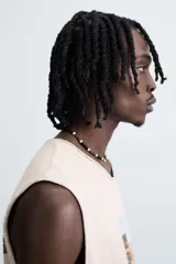 Camiseta tank de punto color beige claro, corte relajado (relaxed fit) y cuello redondo. Presenta un estampado central de un vehículo todoterreno cargado de equipaje conduciendo por una carretera desértica. Los bordes del cuello y las sisas tienen un acabado deshilachado.