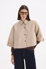 Piloto corto estilo camisa en color beige, con cuello clásico, cierre frontal de botones y mangas anchas con detalle de trabas en los puños.