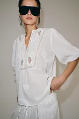 Camisa blanca de algodón con escote en pico, mangas por debajo del codo y detalles de bordados y encaje a tono en el pecho y las mangas.