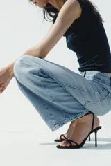 Sandalia de tacón alto fino, color negro, con tiras finas cruzadas en el empeine y un detalle de abalorio rojo y dorado en la unión de las tiras. Posee tira ajustable con hebilla en el tobillo.