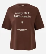 Remera de algodón color marrón oscuro, con cuello redondo y manga corta. Presenta un estampado frontal en blanco con el texto "Sunday Club Palm Paradise", una palmera y una frase motivacional.