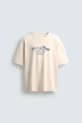 Camiseta de corte boxy fit, color beige con efecto lavado, cuello redondo y manga corta. Presenta un estampado gráfico en azul oscuro en el frente con tipografía estilo vintage.