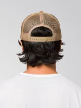 Gorro trucker de color verde militar, con visera curvada marrón claro, panel frontal rígido y red trasera. Cuenta con un parche circular bordado en la parte delantera y cierre ajustable snapback.