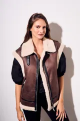 Chaleco oversize estilo gamulán en color marrón, confeccionado en cuero vegano con forro y solapas de corderito sintético en contraste. Posee cierre frontal metálico, bolsillos con cremallera y detalles de correas con hebillas en el cuello.