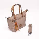 Bandolera tipo shopper color beige con diseño acolchado y pespuntes geométricos. Tiene doble asa de mano en tono tostado y una correa larga regulable y extraíble. Incluye cierre superior, bolsillo frontal con cierre adornado con un llavero de cordón naranja con cuentas y un parche decorativo.