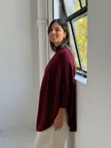 Poncho de punto color bordó con cuello alto y diseño de trenzas verticales en el frente. Presenta mangas tres cuartos con puños acanalados y un corte holgado con ruedo redondeado.