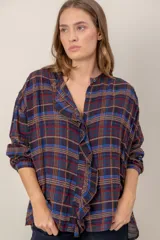 Camisa de corte holgado con estampado de cuadros escoceses en tonos azul, rojo y marrón. Presenta cuello mao, cierre frontal con botones y un detalle de volado vertical a lo largo de la cartera. Mangas largas con puños abotonados.