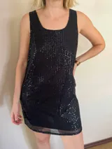 Vestido corto negro de lentejuelas, sin mangas y con escote redondeado. Presenta un ruedo con detalle de tela de red o tul transparente.