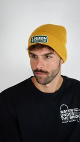 Gorro tipo beanie de tejido de punto color amarillo, con parche rectangular bordado en el frente que incluye el logo y nombre de la marca Duxon.