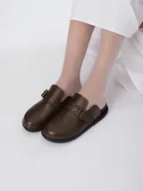 Mules de cuero marrón con textura granulada, punta redondeada y tira ajustable con hebilla metálica y detalles de ojalillos.