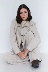 Chaqueta de gabardina color beige con costuras a la vista, cuello camisero, cierre frontal con botones oscuros y mangas largas con puños rectos.