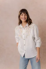 Camisa de denim azul con bordados florales en tonos amarillos y celestes sobre los hombros y el pecho. Presenta cuello clásico, cierre frontal con botones y mangas largas con puños.
