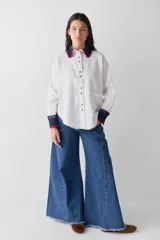 Camisa de lino blanco con cuello y puños tejidos a mano en crochet de color azul con borde rojo. Presenta cierre frontal con botones, bolsillo lateral y costuras a la vista en contraste.