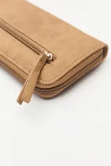 Billetera de formato rectangular con cierre perimetral, confeccionada en material símil cuero de textura suave. Presenta un aplique metálico circular con logo grabado en el frente y tirador de cierre en el mismo material.
