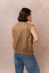 Chaleco de gamuza color marrón con acabado aterciopelado, cuello alto con broche a presión, cierre frontal metálico y bolsillos laterales con cierre.
