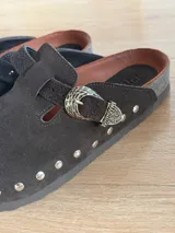 Zueco de gamuza marrón oscuro, forrado en cuero vacuno, con tira ajustable con hebilla metálica decorativa y tachas plateadas alrededor de la suela.