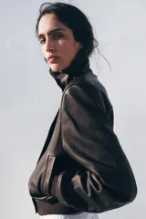 Chaqueta tipo bomber de piel con cuello alto, manga larga con hombreras y puños elásticos. Presenta bolsillos delanteros, bajo elástico y cierre frontal con cremallera y botones ocultos por solapa.