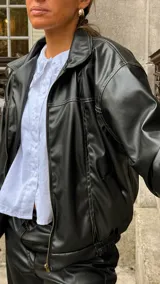 Campera de cuero sintético color negro, con cuello camisero, cierre frontal y costuras visibles en contraste. Presenta un diseño holgado con puños elásticos.