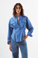 Camisa azul de algodón con finas rayas blancas verticales, cuello mao, manga larga y detalle de pinzas y botones en la zona de la cintura. Posee dos bolsillos de parche con tapa en el pecho.