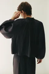 Blusa oversize corta color negro, confeccionada en tejido brillante. Presenta cuello nido de abeja con frunces y escote en V profundo, mangas globo largas de corte raglán y puños elásticos.