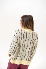Buzo de punto tipo polo en tono amarillo claro con diseño de ondas a rayas en tonos marrón y celeste. Presenta cuello clásico, cierre frontal parcial con botones y silueta relajada con mangas largas.