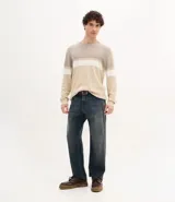 Suéter de punto con cuello redondo y manga larga, diseñado con un patrón de bloques de color en tonos beige, blanco y gris. Presenta un corte regular que se ajusta de forma anatómica al cuerpo.