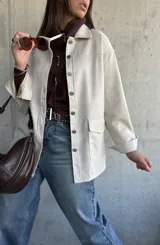 Chaqueta tipo sobrecamisa color beige, con cierre frontal de botones metálicos, cuello clásico y bolsillos frontales. Presenta un diseño de corte recto y mangas largas con puños abotonados.