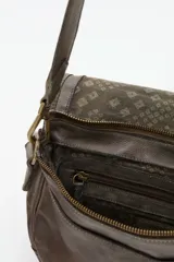 Cartera tipo bandolera de cuero vacuno color gris con diseño redondeado. Presenta una solapa frontal decorada con tachas metálicas en tonos dorados y plateados formando un patrón ornamental, incluyendo un aplique central circular grabado.