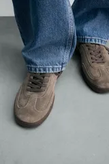 Zapatilla de estilo urbano confeccionada en piel con acabado de serraje en color marrón. Presenta cierre mediante cordones al tono, detalle de pespuntes en el empeine y suela de goma a tono.