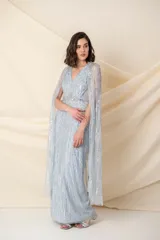 Vestido largo de tul en color celeste pastel, con escote en V y breteles anchos. Presenta un diseño bordado con lentejuelas y pedrería en líneas verticales y una capa larga integrada que nace desde los hombros.
