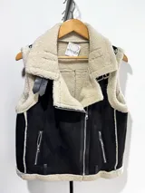 Chaleco estilo gamulán de gamuza color marrón con interior y bordes de corderito sintético color crema. Presenta cierre frontal metálico, bolsillos laterales con cierre y detalles de tiras de cuero sintético en la cintura.