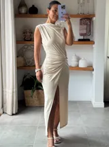 Vestido largo color beige, sin mangas, con escote redondo y detalle de frunces laterales en la cintura que crean un efecto drapeado y una abertura frontal en la falda.