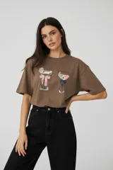 Remera de algodón color marrón con cuello redondo y mangas cortas. Presenta un estampado frontal con ilustraciones de dos copas de cóctel decoradas con lazos.