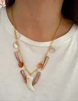 Collar de cadena dorada con múltiples dijes colgantes en tonos rosados, incluyendo piezas de nácar y un dije central en forma de colmillo color crudo, acompañado por un dije circular con diseño de árbol de la vida.