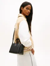 Cartera tote de poliuretano sintético liso color negro de la marca Tommy Jeans, con dos asas superiores y correa ajustable. Presenta cierre de cremallera, compartimento principal e insignia metálica de la marca en la parte delantera. Incluye un adorno colgante extraíble con cordón azul trenzado, dije en forma de corazón y herrajes plateados.